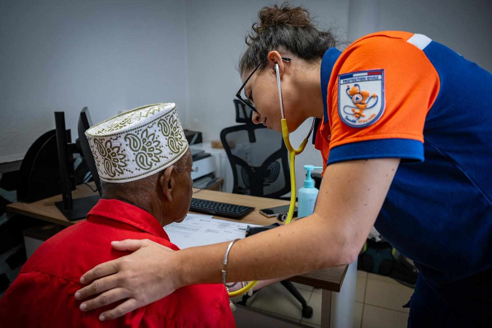 Une médecin de la Protection Civile Santé Mayotte ausculte au stéthoscope un homme âgé assis dans un bureau de consultation.